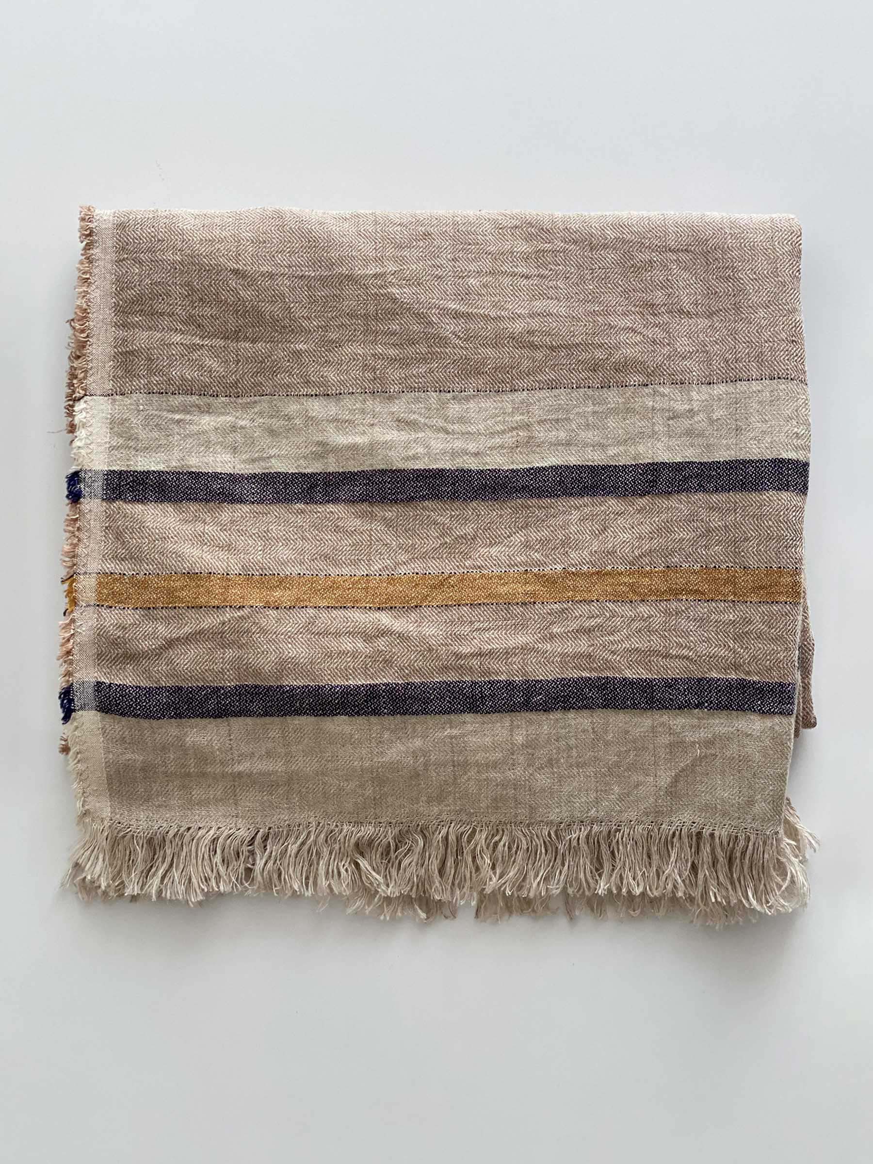 Arlo woven throw in sand dune colour with contrasting stripes folded on a white surface showing French flax linen texture herringbone weave and fringed edges